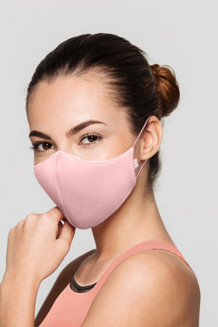 A model is wearing a B-Safe Adult Face Mask in a soft pink color, showcasing its snug fit and stylish design. The mask features adjustable ear loops and covers the nose and mouth, complementing the models casual outfit.