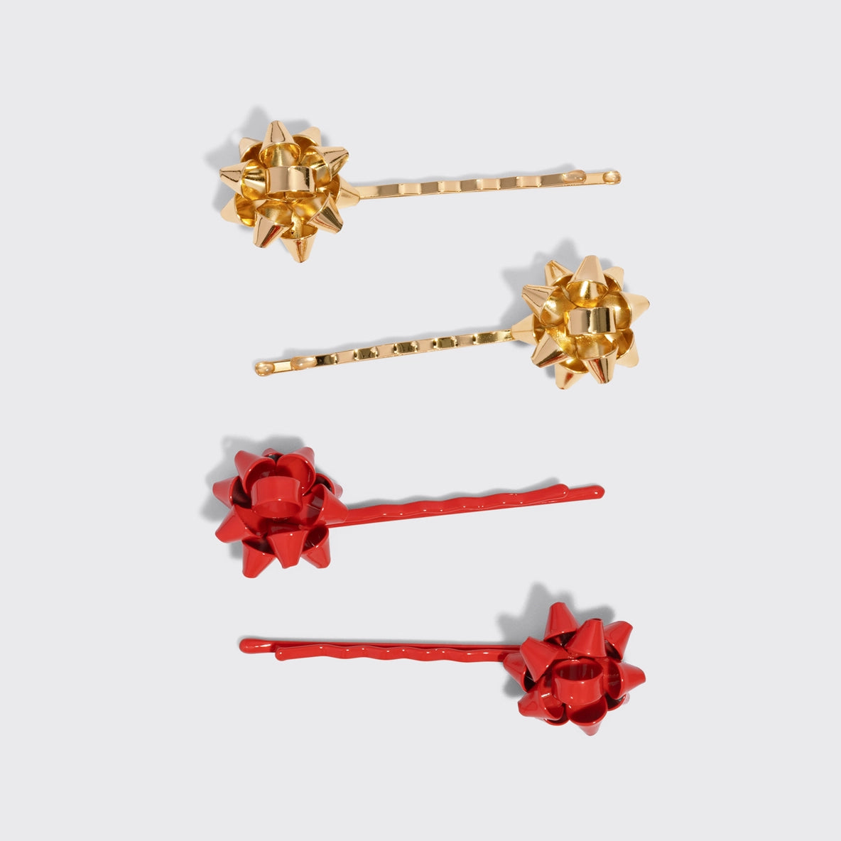 Four bobby pins from the Holiday Gift Bow Bobby Pins 4pc Set, featuring two gold pins with shiny gift bow decorations and two red pins with matching bow designs, are displayed against a light gray background.