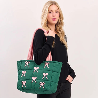 A model poses with a green quilted tote bag featuring pink bow embroidery accents. The tote has a spacious design and is carried casually over her shoulder, showcasing its stylish and functional nature.