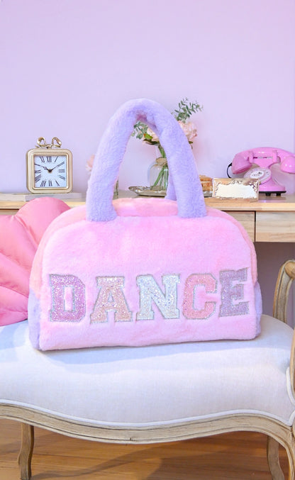 A pastel color-blocked plush duffle bag features a vibrant pink and purple design with glittery varsity-letter patches spelling DANCE prominently on the front.