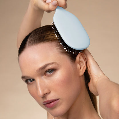 A woman with long, straight hair is brushing her hair with a light blue Smooth & Polish Brush. She is holding the brush at the roots of her hair, and her expression is focused, highlighting the brushs gentle detangling capabilities.
