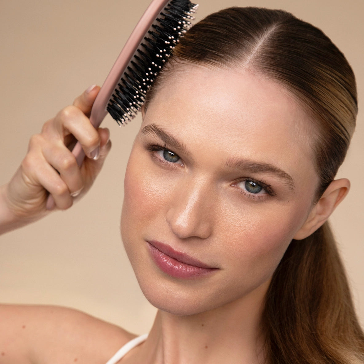 A woman with long, straight hair is using a Smooth & Polish Brush with a light pink handle. The brush features dual bristles, with vegan boar bristles on one side and nylon bristles on the other, and she is lifting the brush towards.