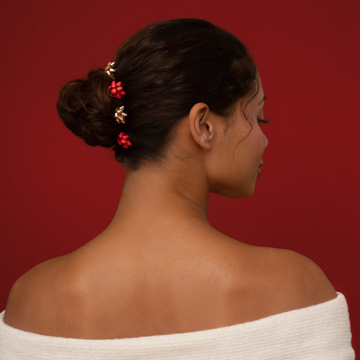 A woman with dark, curly hair is styled in a low bun, adorned with four decorative bobby pins featuring festive red and gold gift bow details.