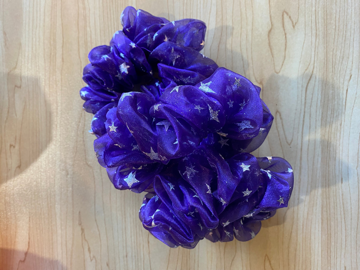 A vibrant purple scrunchie featuring a shimmering star pattern is displayed on a wooden surface. The handmade accessory has a soft, flowing texture, showcasing the premium fabric and unique design.