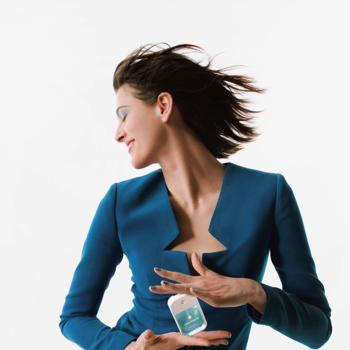 A woman with flowing hair is captured in profile, wearing a vibrant blue blazer. She is holding a frosted mint-colored bottle of hand sanitizer, showcasing its sleek design against a stark white background, exuding a refreshing.