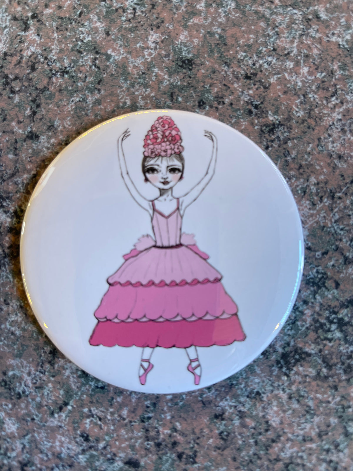 A round pocket mirror featuring a whimsical illustration of a ballerina dressed in a pink gown with layered ruffles.