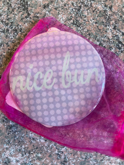 A round pocket mirror with a pastel purple background featuring a subtle pattern. The mirror is partially covered by a sheer pink pouch, and the phrase nice bun is written in a playful, white font across the front.