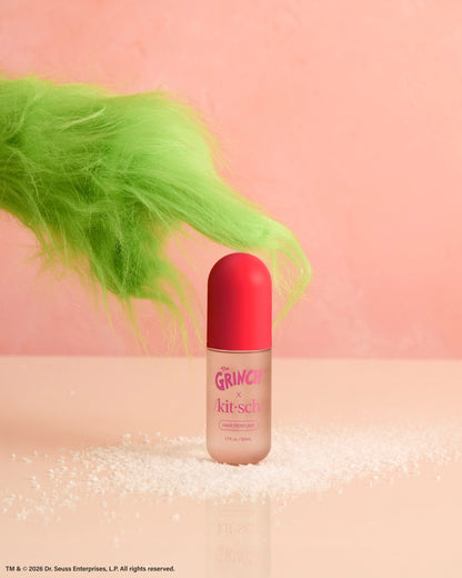 A bottle of The Grinch x Kitsch Cherry Cheer Hair Perfume sits on a light pink surface, surrounded by a sprinkle of white crystals.
