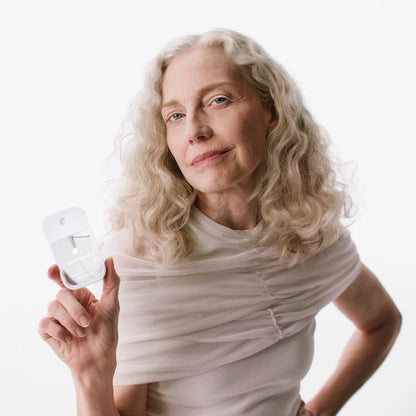 A woman with long, wavy gray hair poses confidently while holding a clear bottle of Unscented Power Mist Hydrating Hand Sanitizer.
