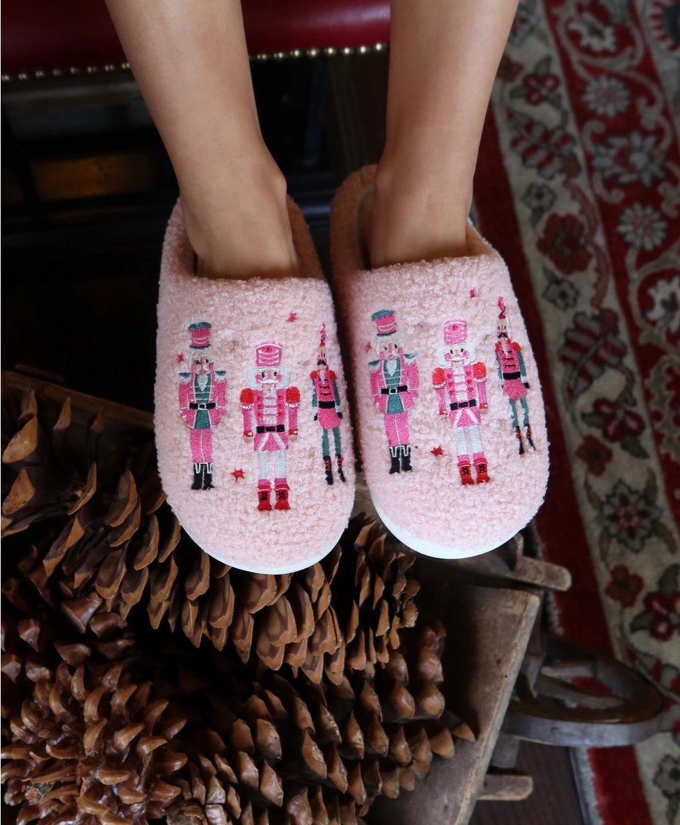 A pair of Living Royal Adult Nutcracker Slippers in soft pink is showcased on a persons feet, featuring colorful nutcracker designs embroidered on the front.