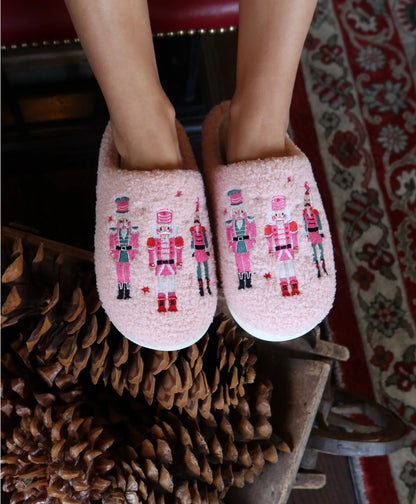 A pair of Living Royal Adult Nutcracker Slippers in soft pink is showcased on a persons feet, featuring colorful nutcracker designs embroidered on the front.