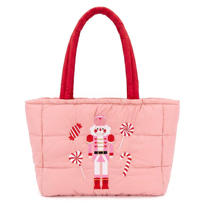 A festive pink quilted tote bag features an embroidered nutcracker surrounded by candy cane designs. The bag has red shoulder handles and a spacious main compartment, perfect for carrying holiday essentials.