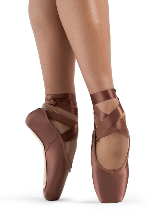 A dancers feet are shown in the 1142W (Ava) pointe shoes in mocha color. The shoes feature a broad inner toe box, satin finish, and crisscross ribbons, highlighting their elegant design.