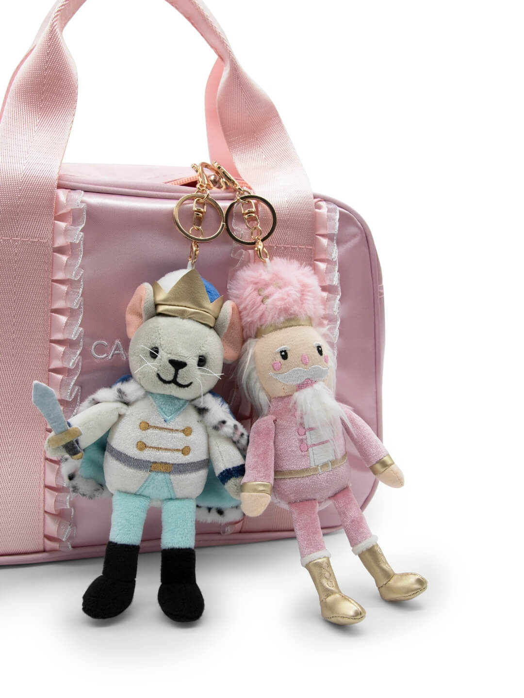 A plush Mouse King keychain, featuring a crown and a blue uniform with a flowing cape, hangs alongside a pink Nutcracker keychain on a pink bag.
