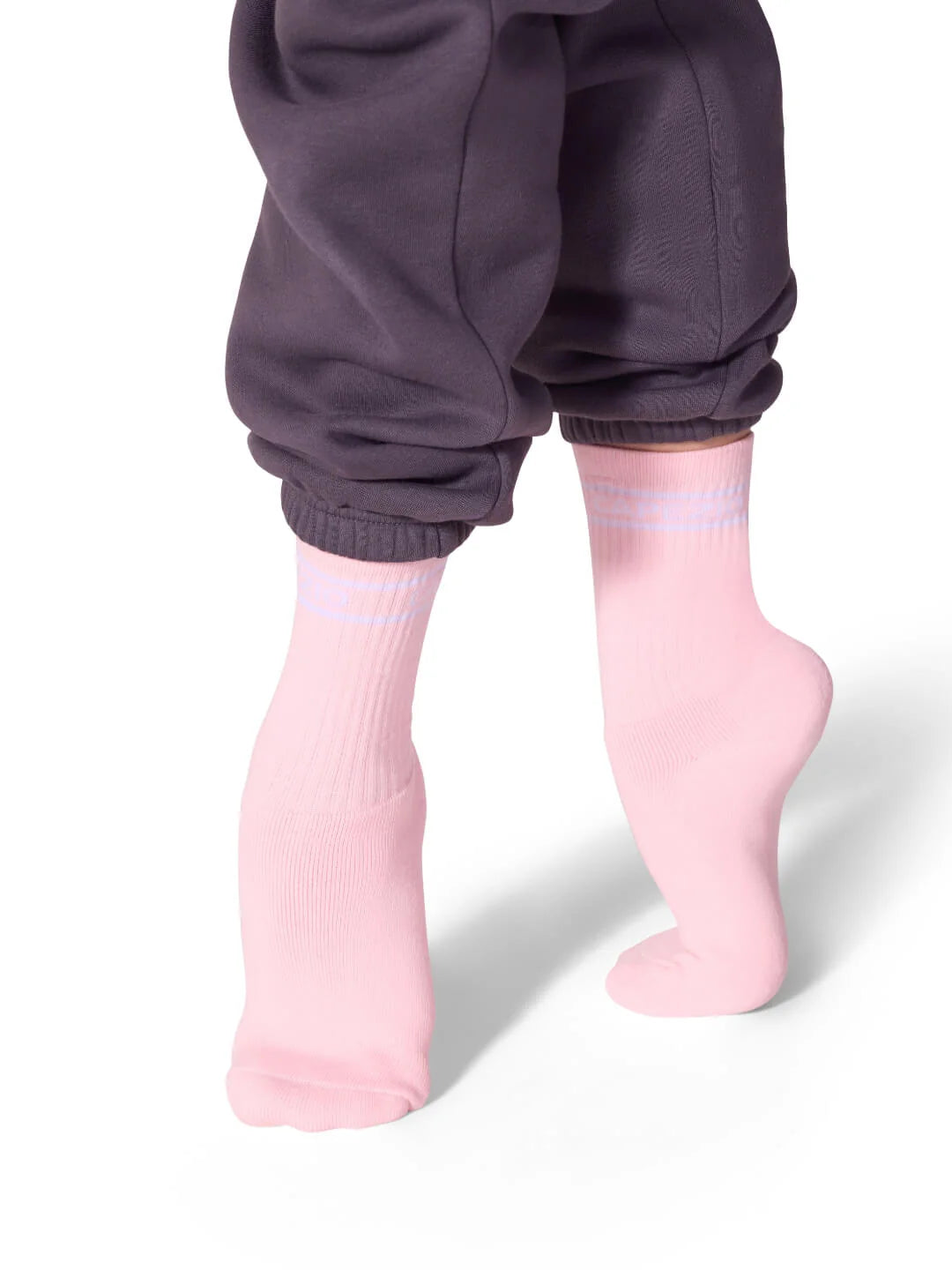 A pair of unisex Varsity Crew Socks in a soft pink color is worn on feet that are positioned slightly to the side, showcasing the socks ribbed texture and subtle design.