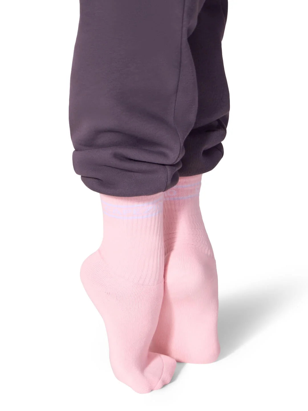 A person is standing on tiptoes, wearing pale pink Varsity Crew Socks with subtle blue stripes near the top. The socks are paired with dark purple jogger pants, creating a stylish contrast that highlights the vintage aesthetic of the socks.