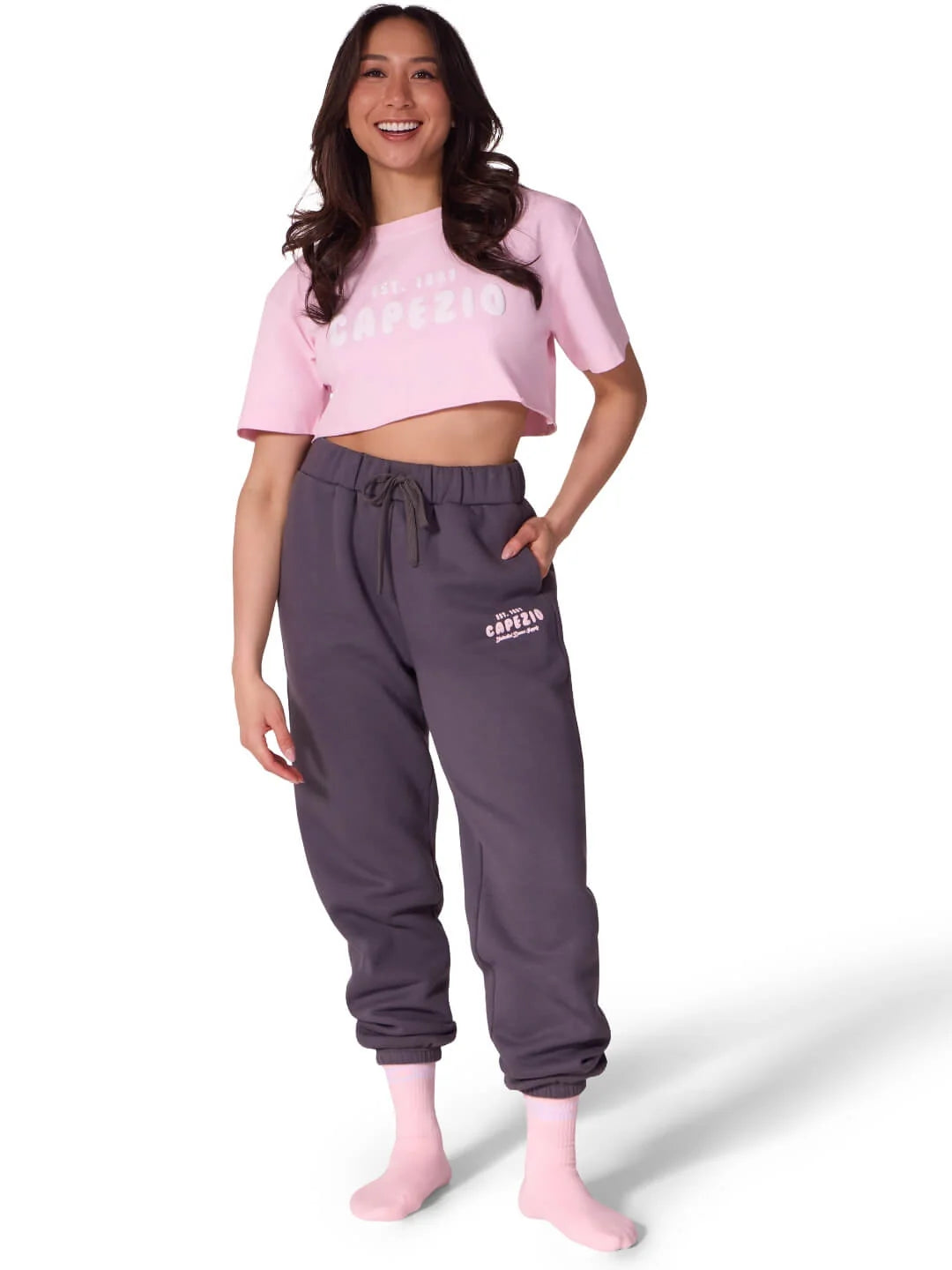 A woman stands smiling, wearing a pink cropped t-shirt with the word Capezio printed in white, paired with dark gray sweatpants featuring the brands logo.