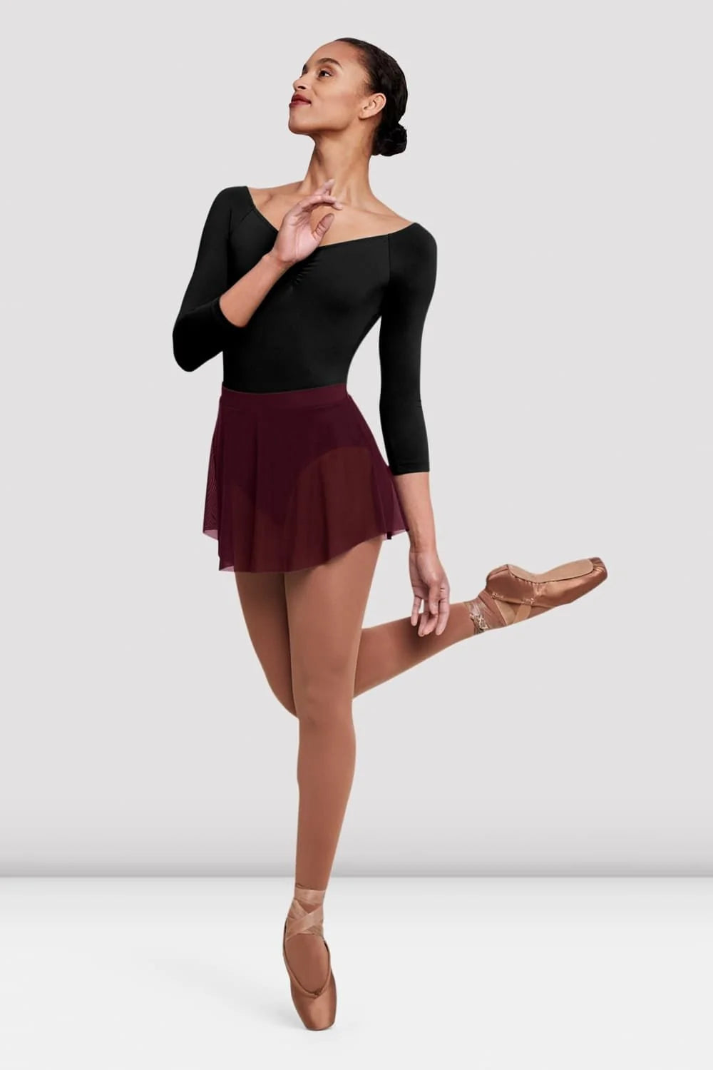 A dancer poses gracefully in a black long-sleeve leotard, paired with a short maroon Ladies Jaye Mesh Skirt by the brand.