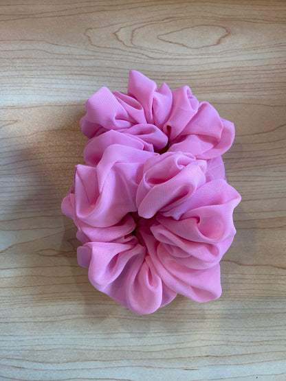 A handmade scrunchie in a soft pink color is displayed on a light wooden surface. The scrunchie features a ruffled design, showcasing its premium fabric and unique texture, making it a stylish accessory for hair.