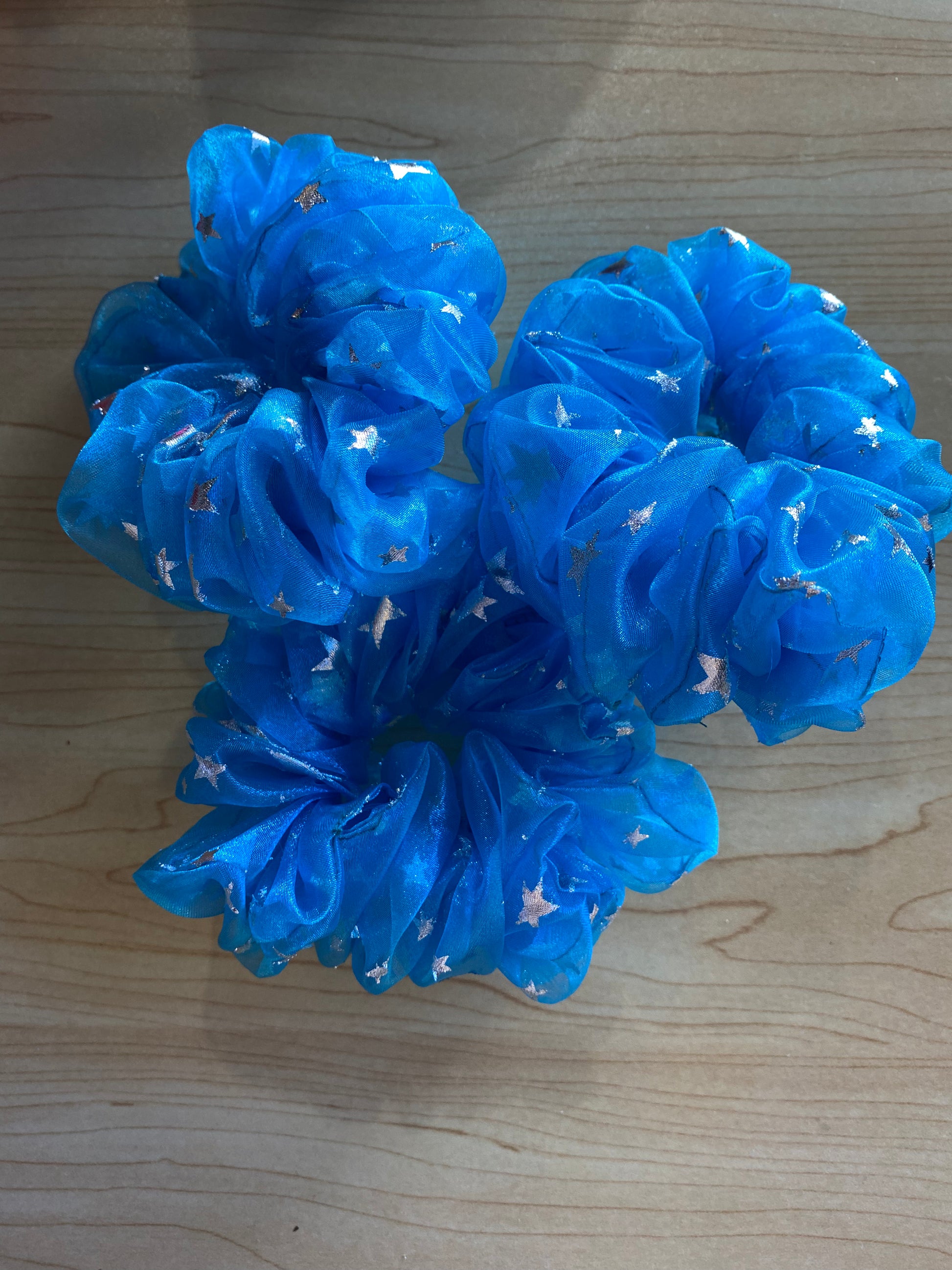 Bright blue scrunchies with a sheer, shimmering fabric adorned with small gold star patterns are arranged together on a wooden surface.