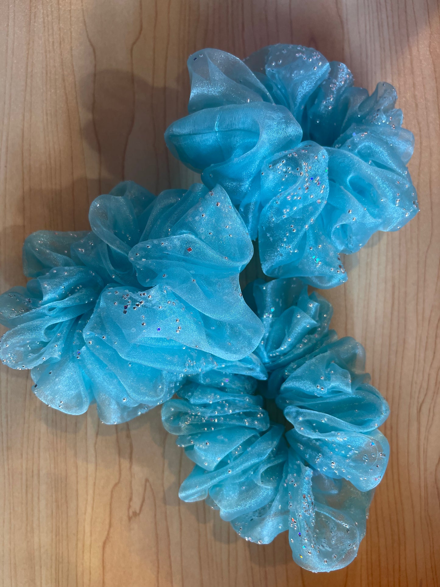Three handmade scrunchies in a vibrant turquoise color are displayed on a wooden surface. Each scrunchy features a sheer, sparkly fabric that shimmers with silver glitter, creating an eye-catching effect.