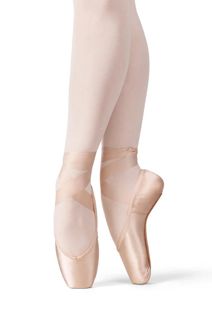 A dancer is poised in an elegant ballet pose, showcasing the Akoya Pointe Shoe in a soft, peachy hue. The shoe features a tapered box and is worn with light, sheer tights, highlighting its streamlined design and flexible shank,.
