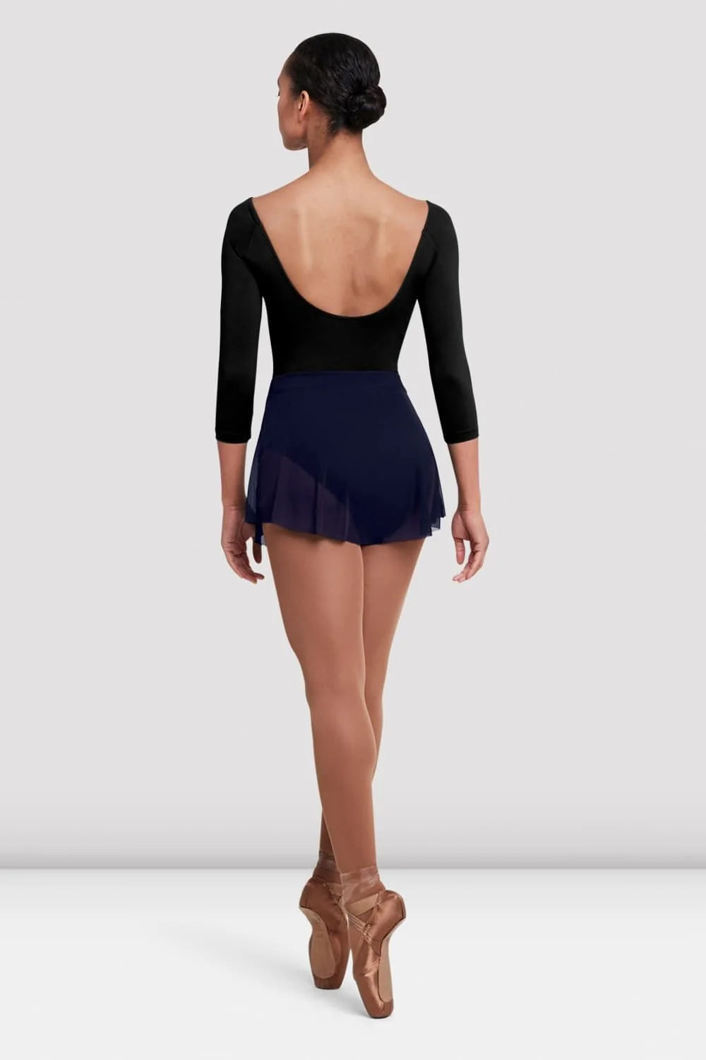 A dancer is seen from the back, wearing a black long-sleeve leotard paired with the Ladies Jaye Mesh Skirt by the brand. The skirt is short and navy blue, featuring a soft, flowing mesh design that accentuates the dancer’s movement.