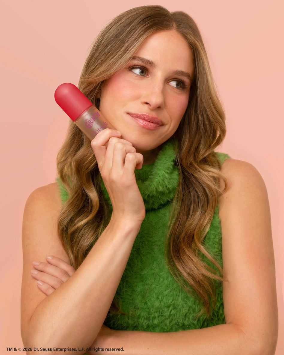 A woman with long, wavy hair is holding The Grinch x Kitsch Cherry Cheer Hair Perfume, which features a clear bottle with a pink cap.