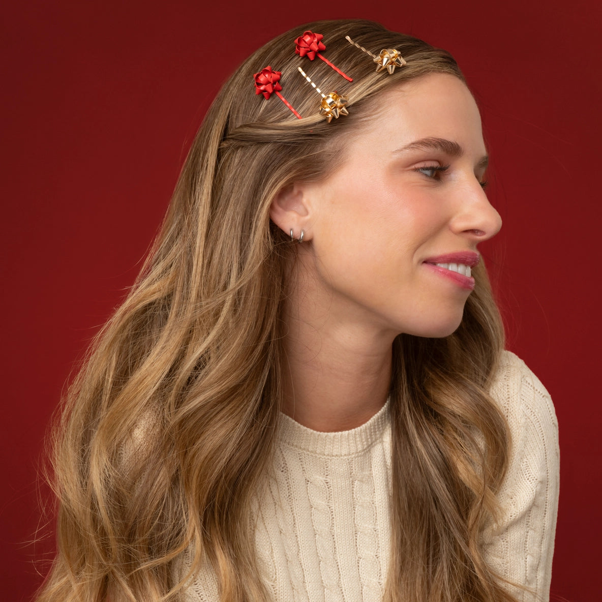 A woman with long, wavy hair is wearing a set of four Holiday Gift Bow Bobby Pins in red and gold. The bobby pins, featuring festive bow designs, are positioned in her hair against a rich red background, complementing her.