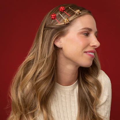 A woman with long, wavy hair is wearing a set of four Holiday Gift Bow Bobby Pins in red and gold. The bobby pins, featuring festive bow designs, are positioned in her hair against a rich red background, complementing her.