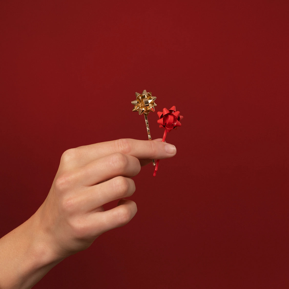 A hand is holding two bobby pins from the Holiday Gift Bow Bobby Pins 4pc Set, featuring a festive red and gold color scheme.