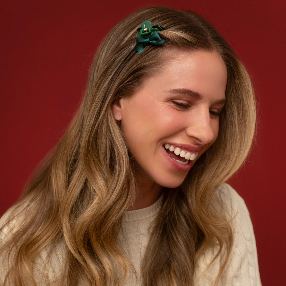 A woman with long, wavy hair is smiling while wearing a green mini claw clip adorned with a small bow, which is positioned in her hair on the left side.