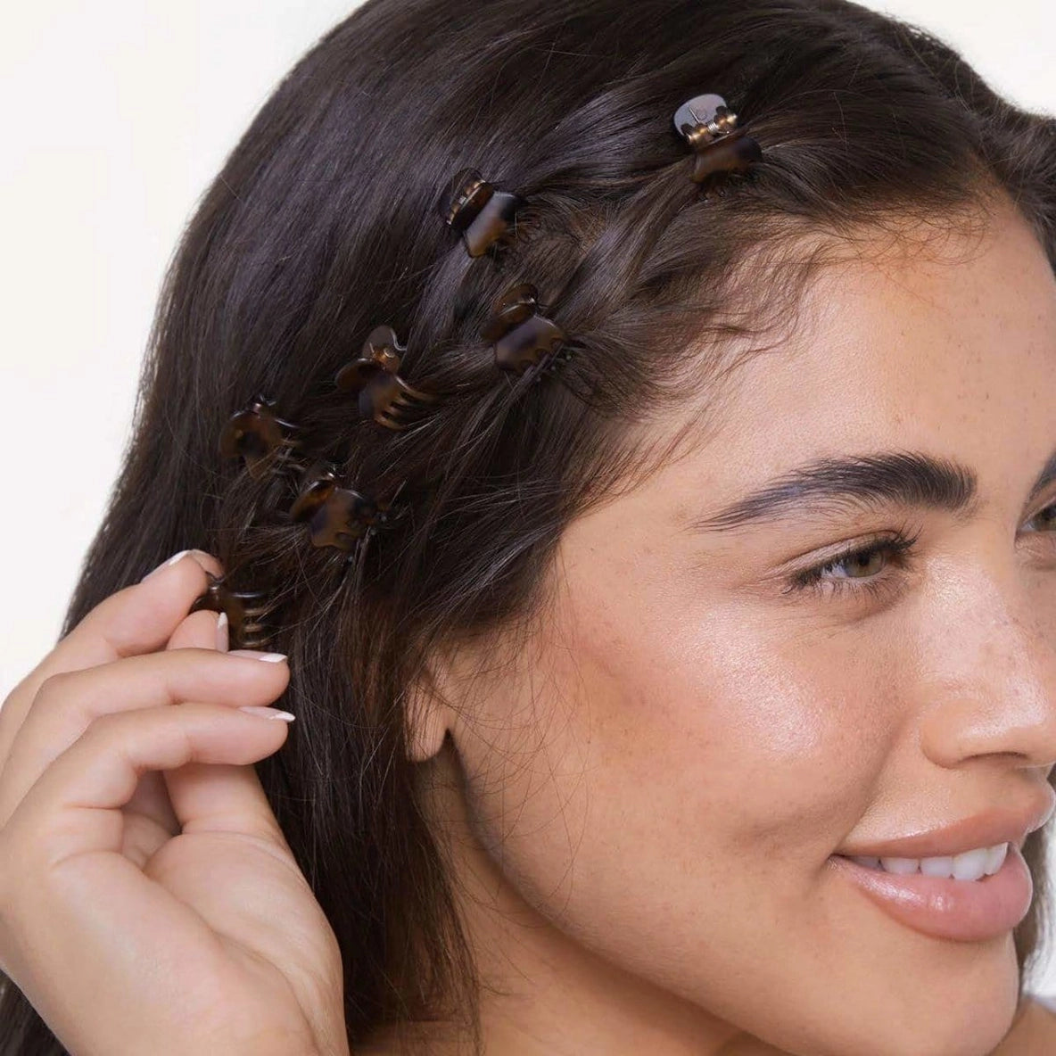 A woman with long, dark hair is smiling while showcasing multiple Kitsch Mini Claw Clips in a tortoiseshell pattern.