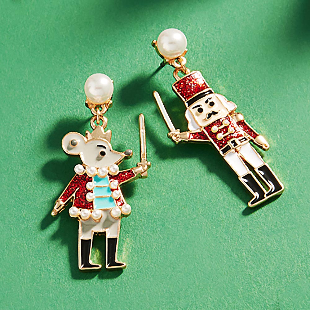 Whimsical enamel Nutcracker and Mouse dangle earrings are showcased against a green background. The earrings feature playful details, with the Mouse holding a sword and wearing a festive outfit, while the Nutcracker stands tall, both.