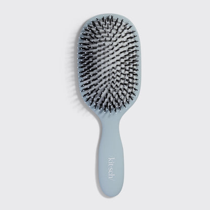 The image features a Kitsch Smooth & Polish Brush in a soft blue color. The brush design includes dual-bristle technology, showcasing both vegan boar bristles for smoothing and nylon bristles for detangling, all set in a comfortable,.