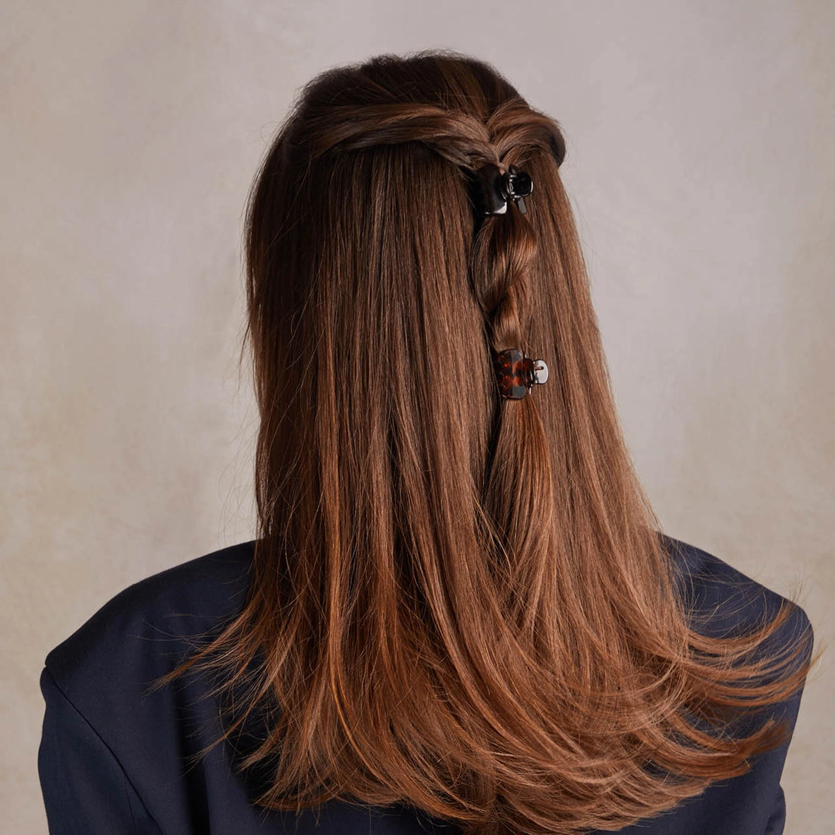A model with long, straight brown hair is seen from the back, featuring a stylish half-up hairstyle. Two mini claw clips from the Recycled Plastic Mini Puffy Claw Clips 6pc Set in black and tortoise colors hold back sections of hair,.