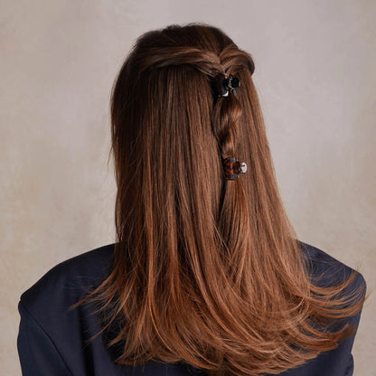 A model with long, straight brown hair is seen from the back, featuring a stylish half-up hairstyle. Two mini claw clips from the Recycled Plastic Mini Puffy Claw Clips 6pc Set in black and tortoise colors hold back sections of hair,.