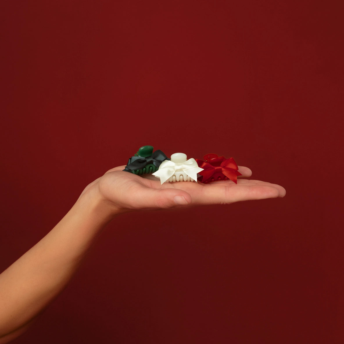 A hand holds a selection of three mini claw clips against a rich red background. The clips include one green, one white, and one red, showcasing a festive holiday theme, perfect for securing hair while adding a pop of color to any.