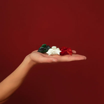 A hand holds a selection of three mini claw clips against a rich red background. The clips include one green, one white, and one red, showcasing a festive holiday theme, perfect for securing hair while adding a pop of color to any.