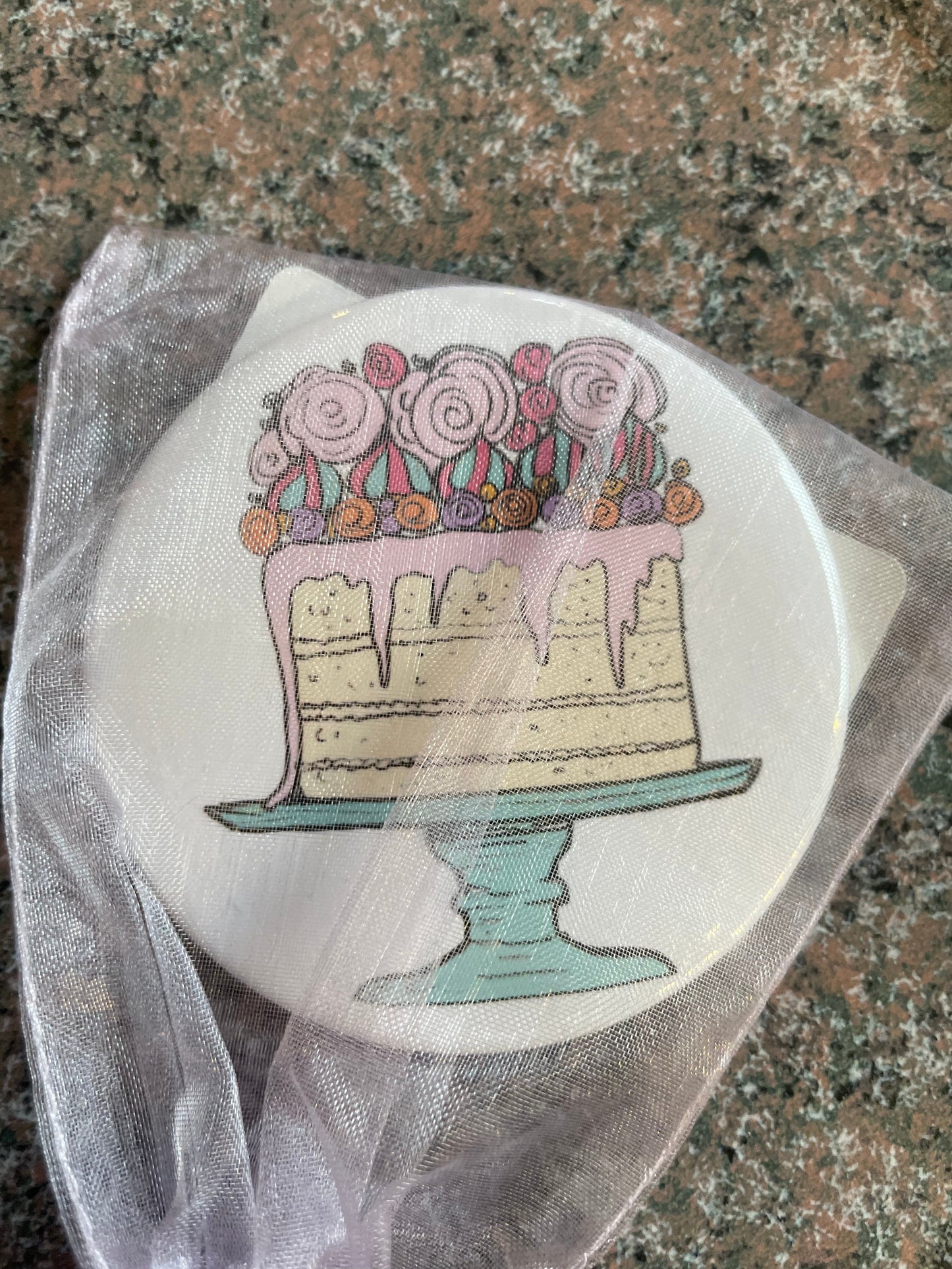 A round pocket mirror featuring a whimsical illustration of a decorated cake on a teal pedestal. The cake is adorned with colorful roses and dripping icing, presented against a light background and wrapped in a sheer, pale ribbon.
