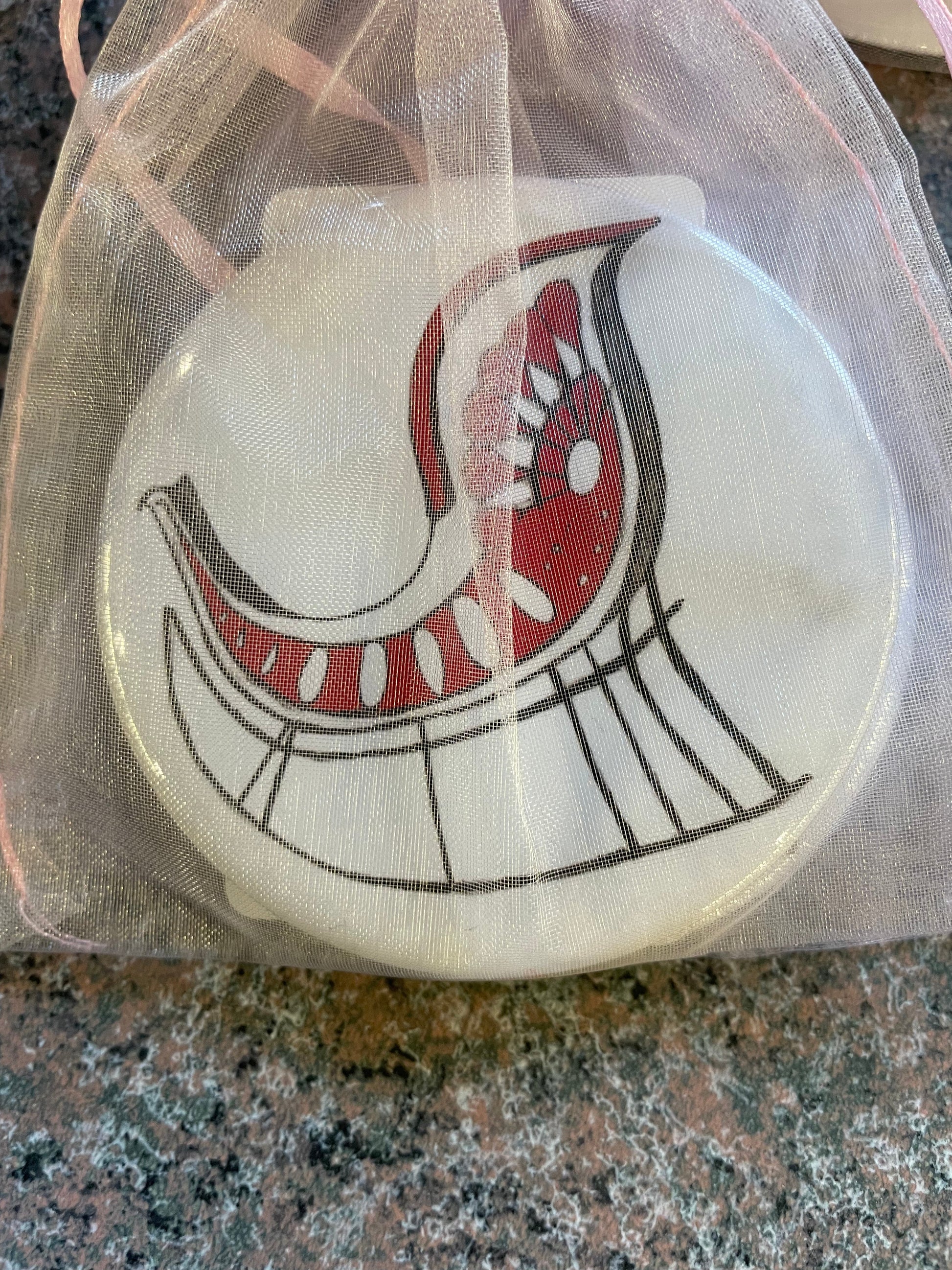 A Nutcracker-themed pocket mirror, featuring a whimsical red and white bird design, is displayed inside a sheer, light-colored drawstring bag.
