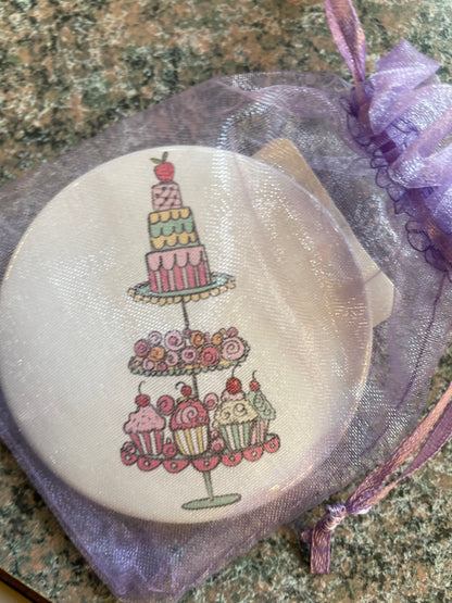 A round pocket mirror featuring whimsical cake and cupcake illustrations is displayed against a textured surface.