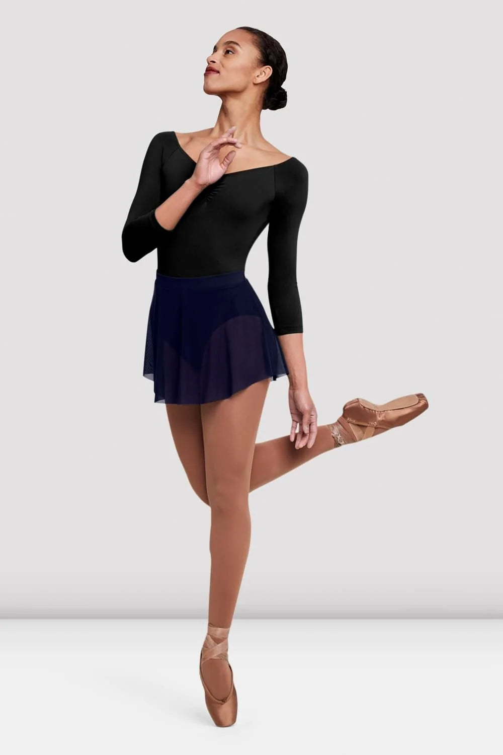 A dancer poses gracefully in a black long-sleeve leotard paired with the Ladies Jaye Mesh Skirt by the brand, featuring a soft, navy mesh fabric.